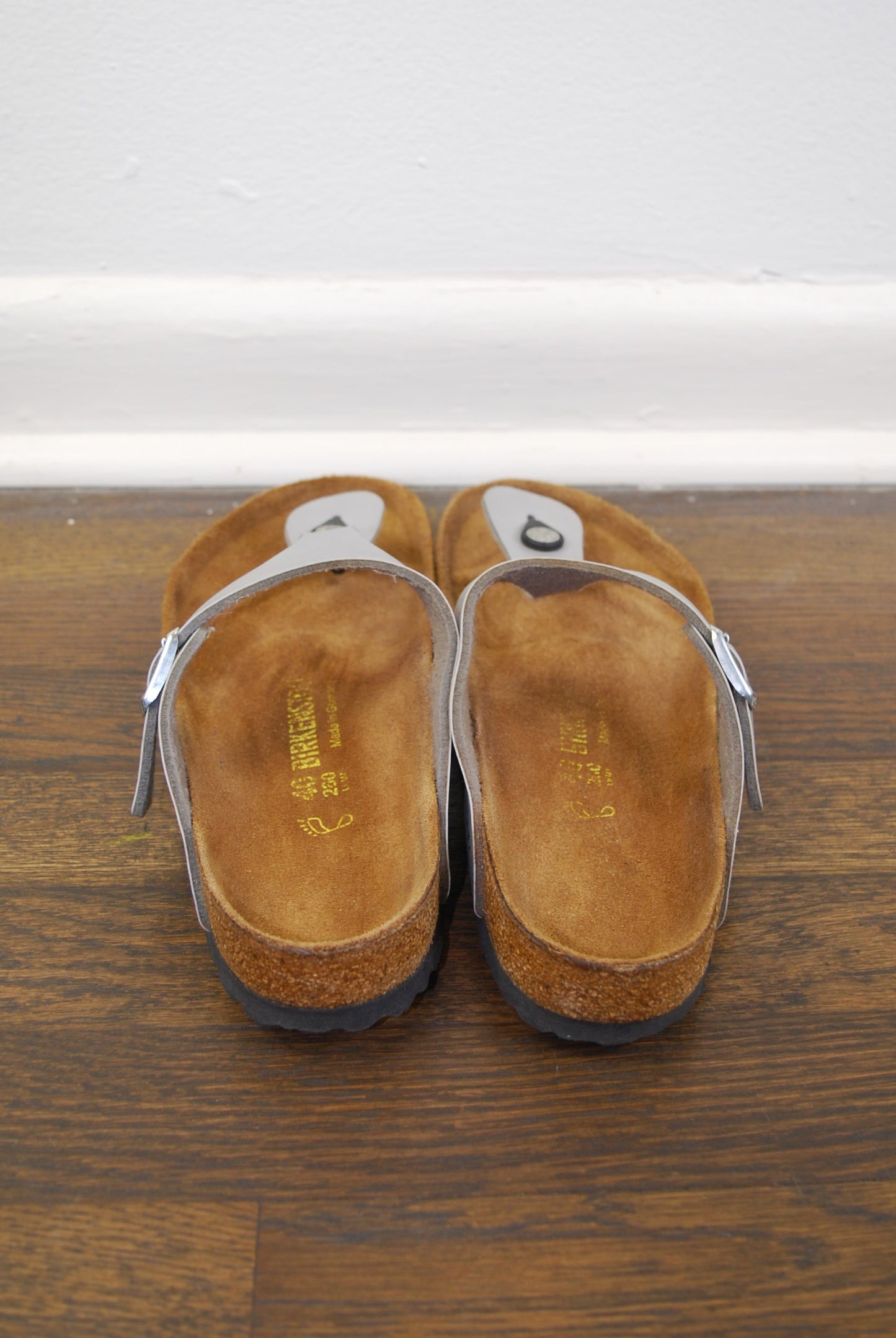 Pair of metallic sandals on a wooden floor with a white wall in the background