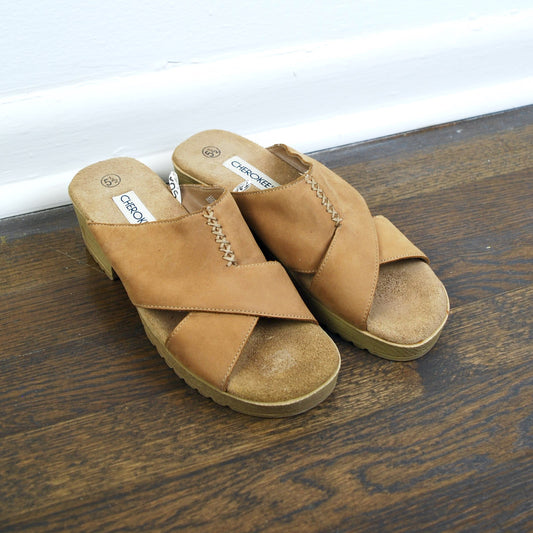 Pair of tan sandals on a wooden floor with a white baseboard.