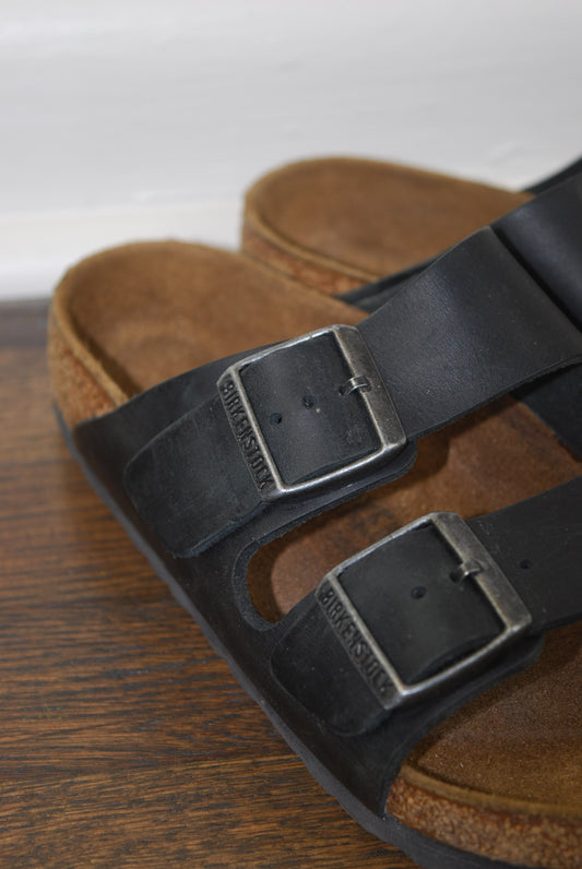 Brown leather sandals with black straps and buckles on a wooden surface.