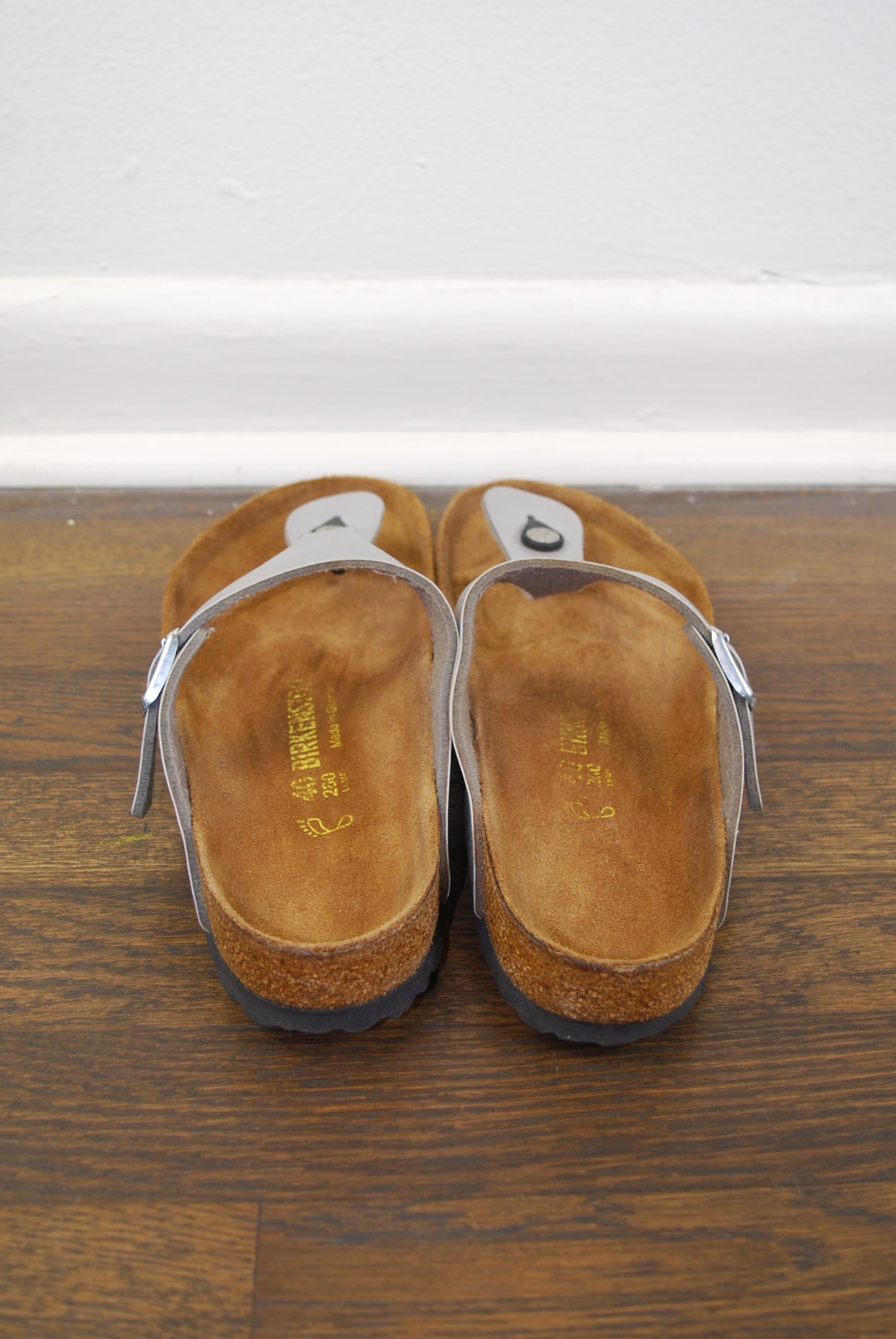 Pair of metallic sandals on a wooden floor with a white wall in the background