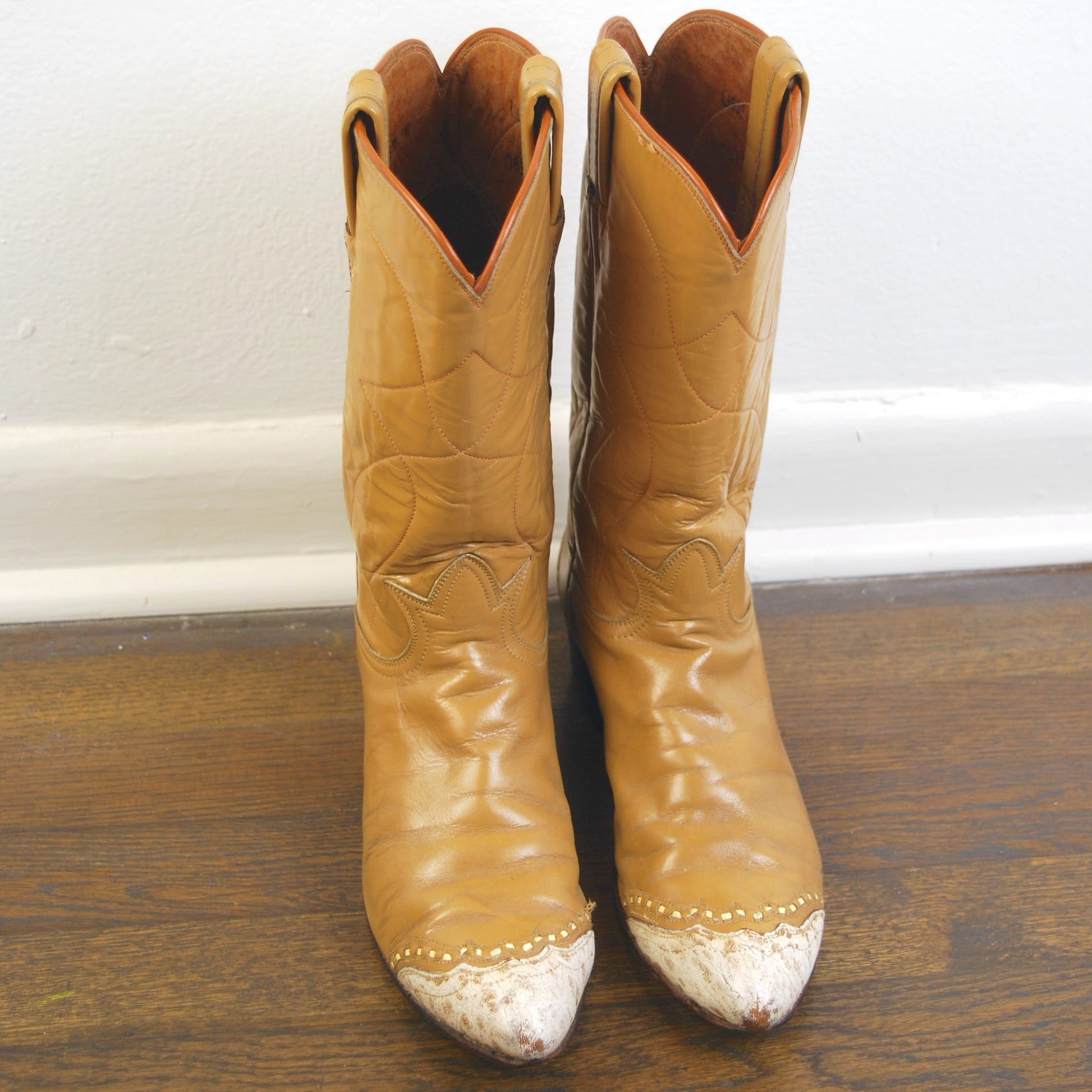 Pair of tan cowboy boots with white soles on a wooden floor.