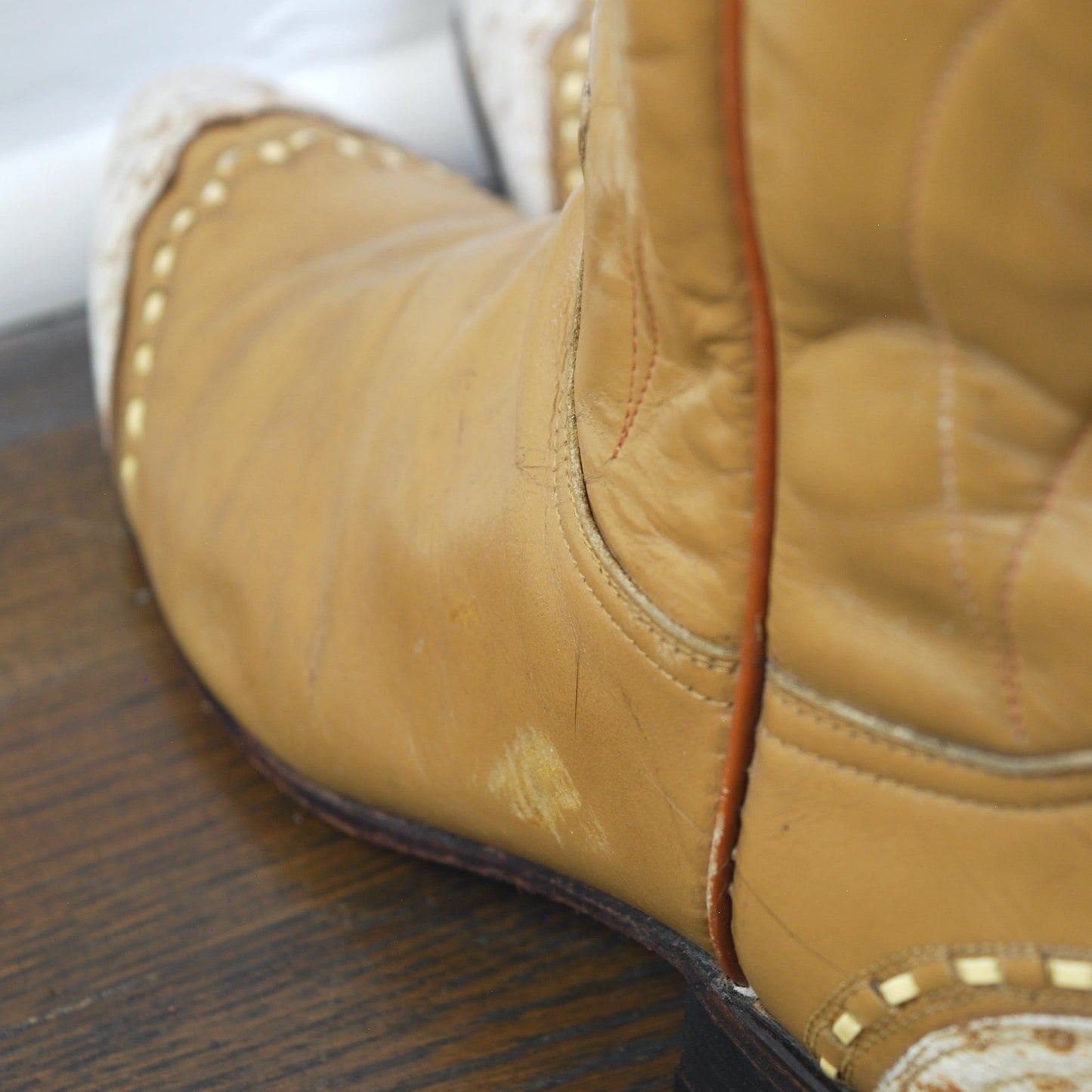 Close-up of a worn brown leather boot with white laces on a wooden floor.