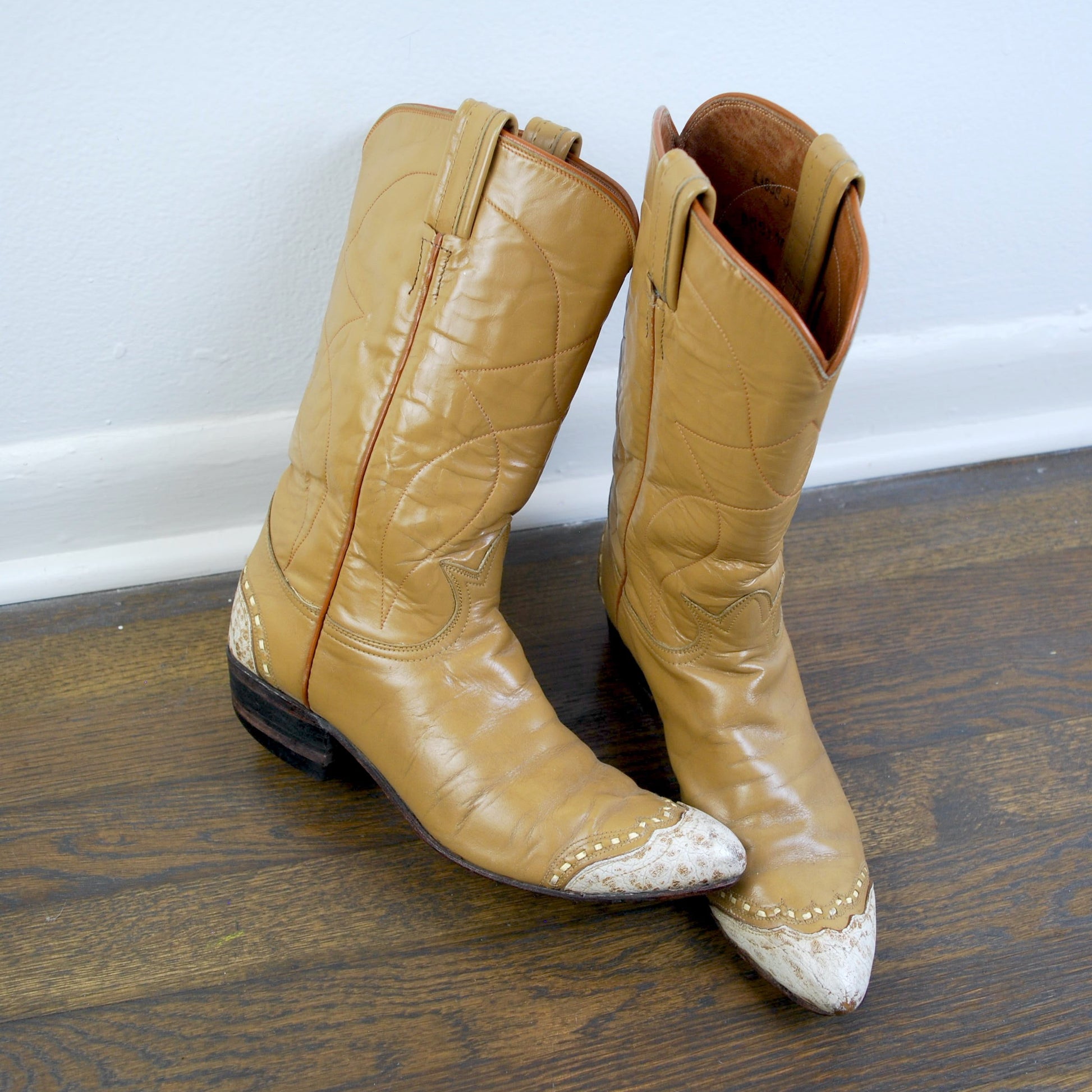 Pair of tan cowboy boots on a wooden floor with a white wall background