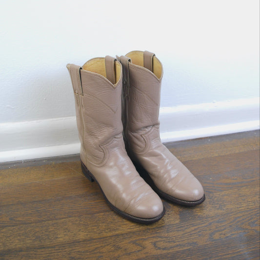 Pair of beige cowboy boots on a wooden floor against a white wall.