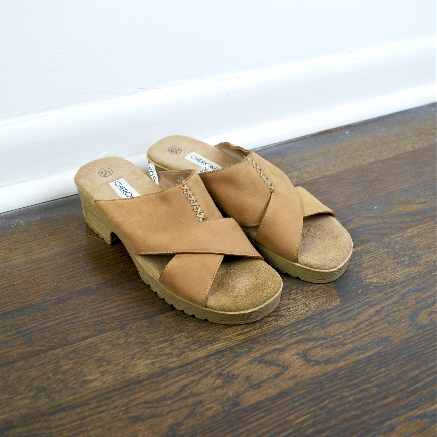 Pair of tan sandals on a wooden floor with a white baseboard.