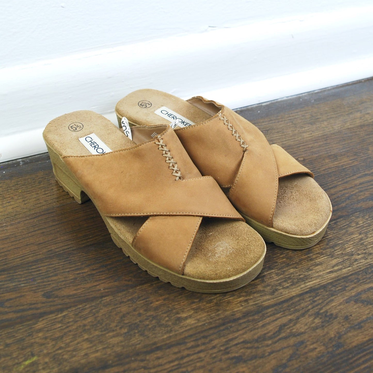 Pair of tan sandals on a wooden floor with a white baseboard.
