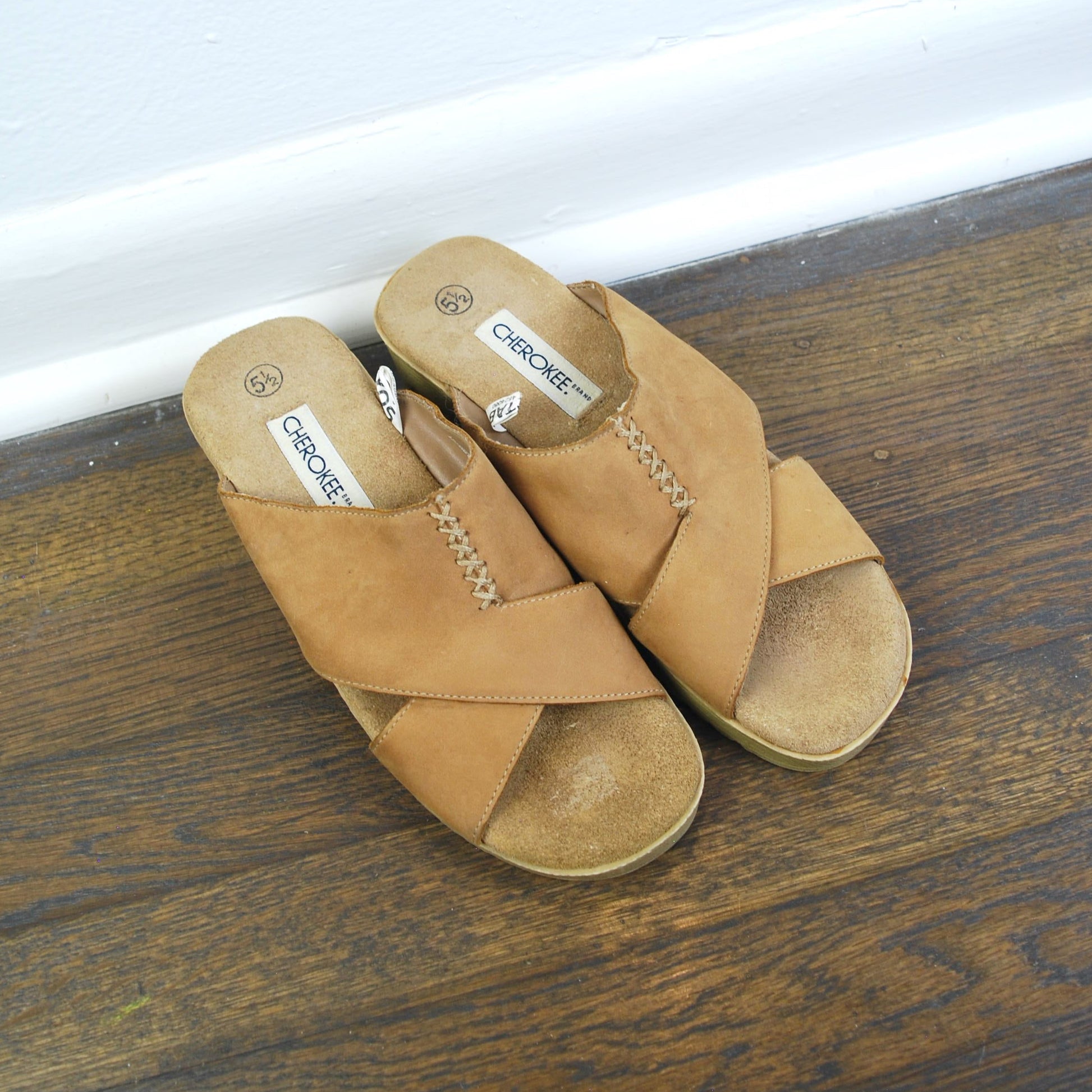Pair of tan sandals on a wooden floor with a white baseboard.