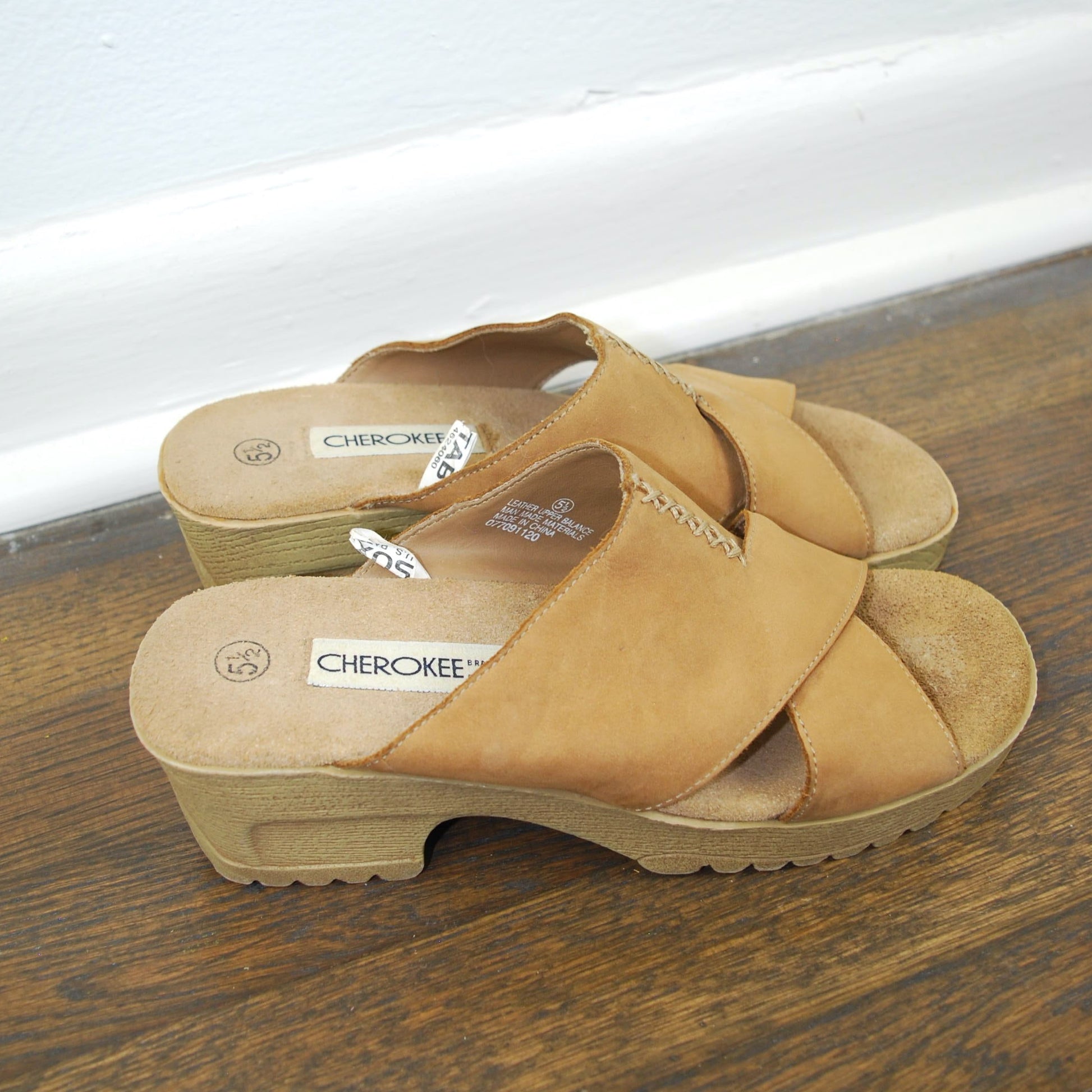 Pair of tan sandals on a wooden floor with a white baseboard in the background.