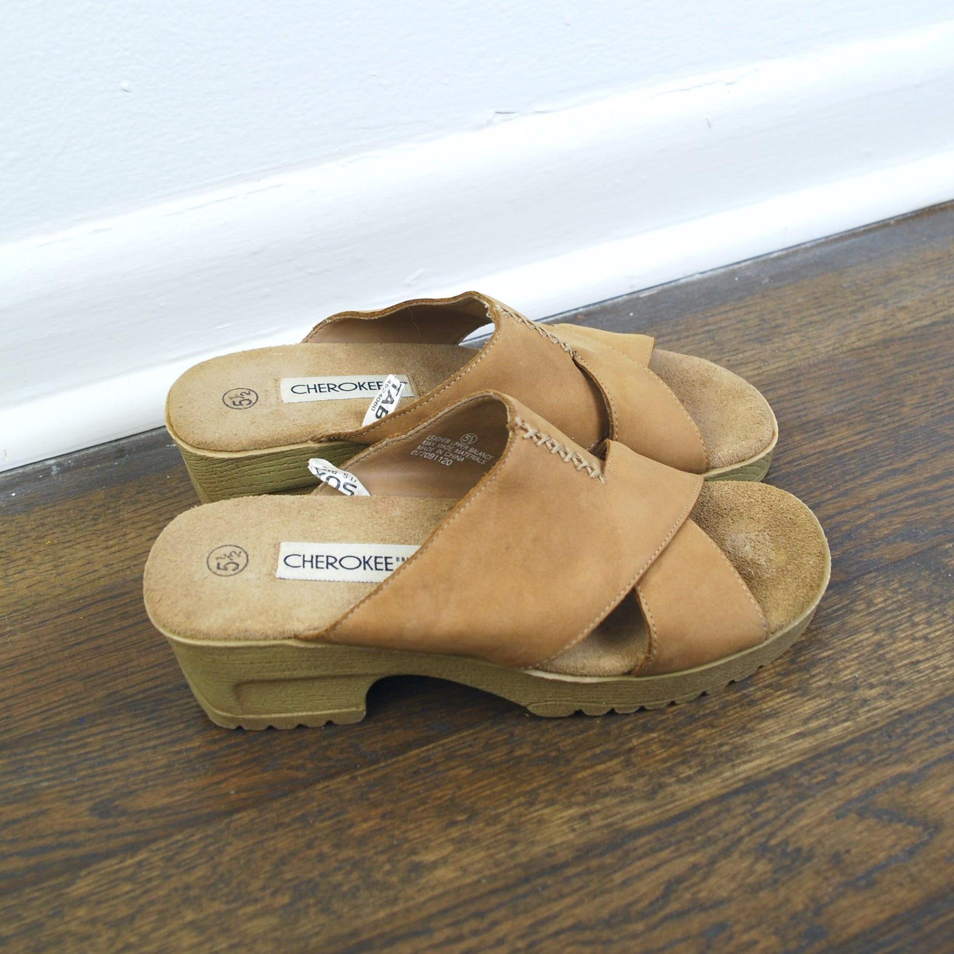 Pair of tan sandals on a wooden floor with a white baseboard in the background.