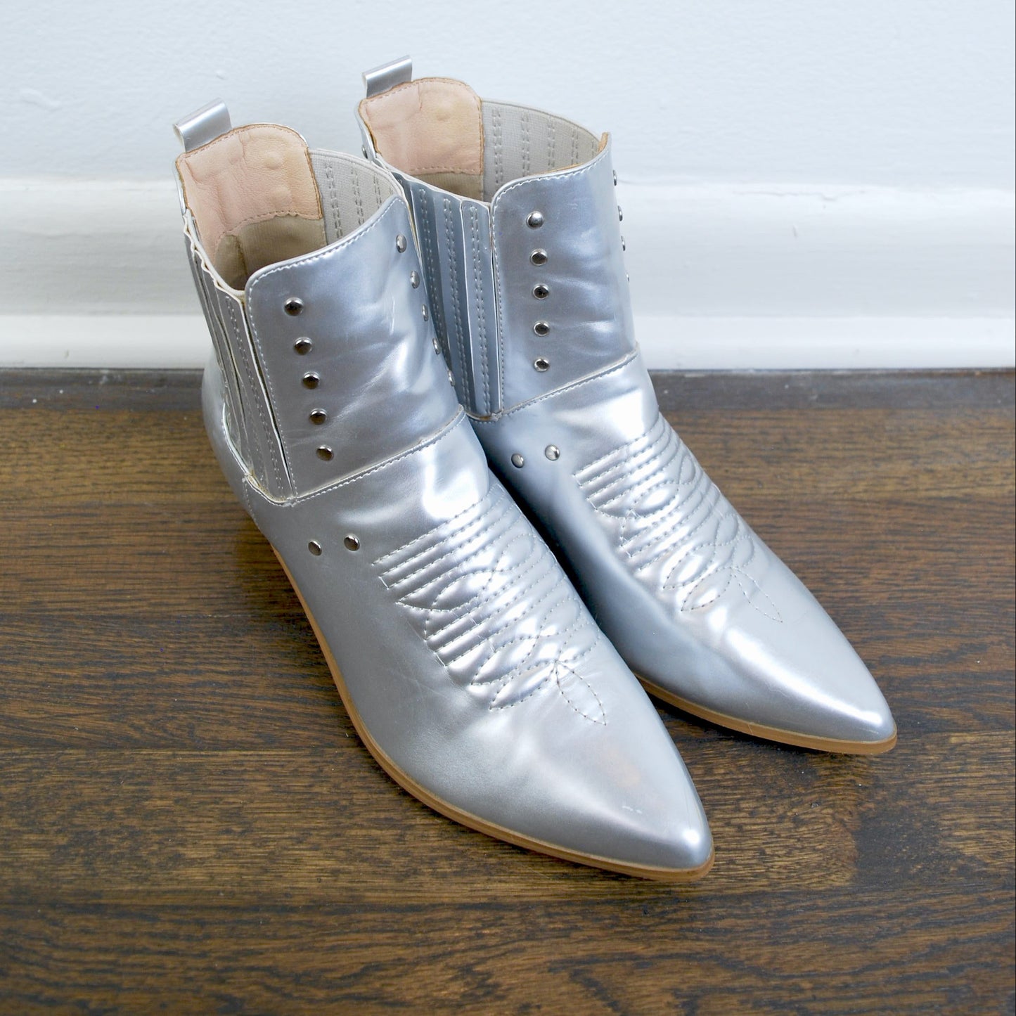 Pair of silver pointed-toe boots on a wooden floor with a light blue wall background.