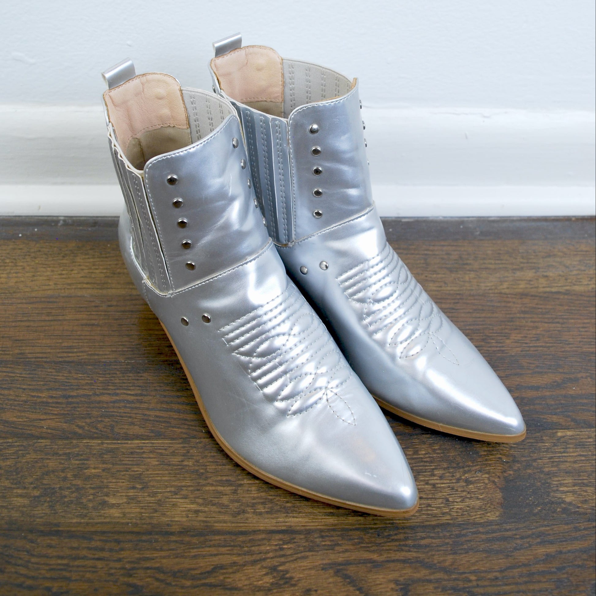 Pair of silver pointed-toe boots on a wooden floor with a light blue wall background.