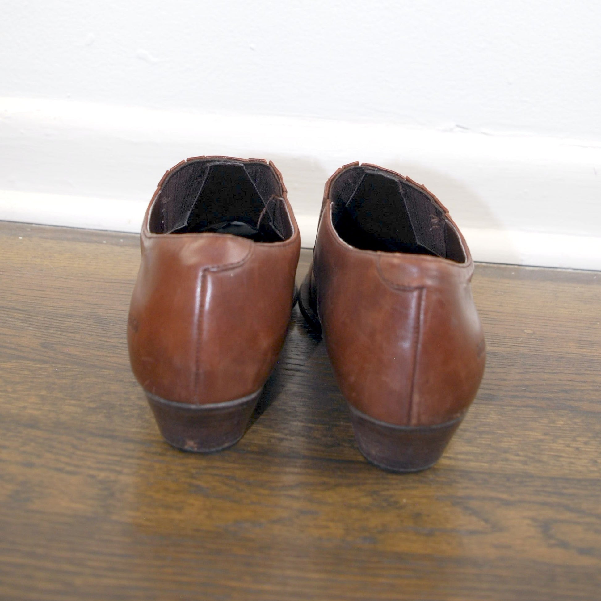 Pair of brown leather shoes on a wooden floor with a white baseboard.
