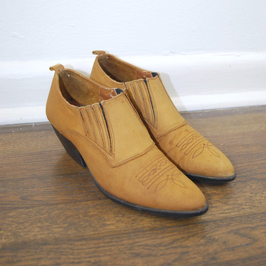 Pair of tan ankle boots on a wooden floor with a white baseboard.