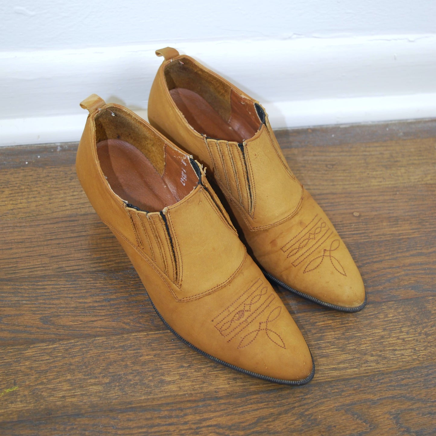 Pair of tan shoes on a wooden floor with a white baseboard in the background