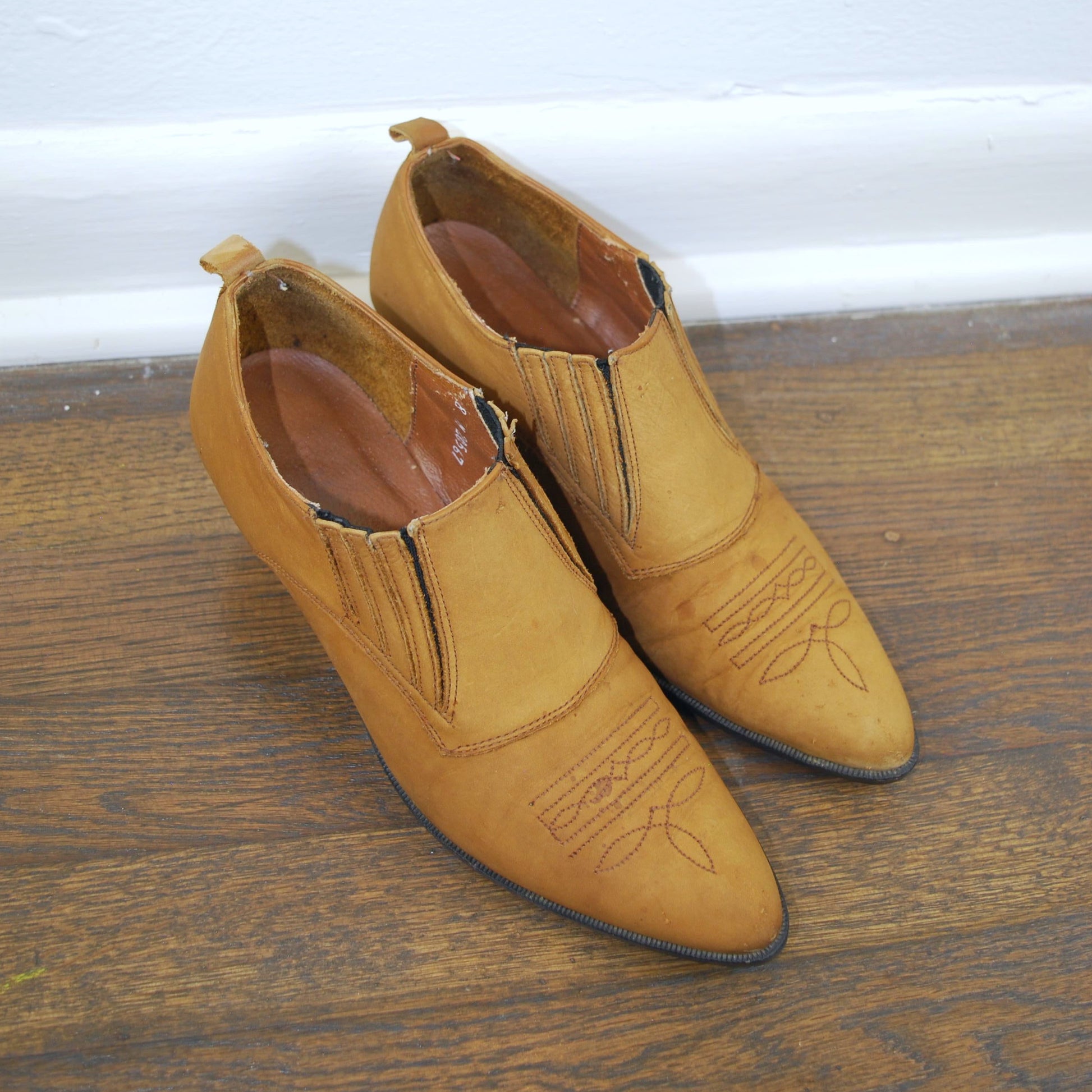 Pair of tan shoes on a wooden floor with a white baseboard in the background