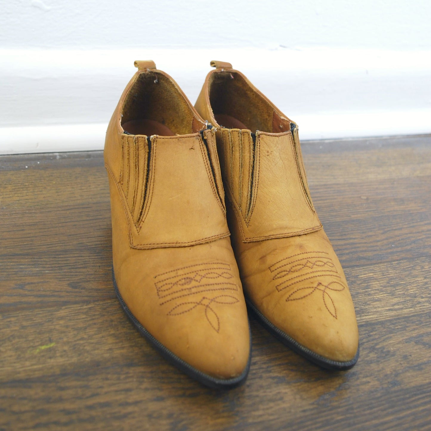 Pair of tan leather boots on a wooden floor with a white wall background