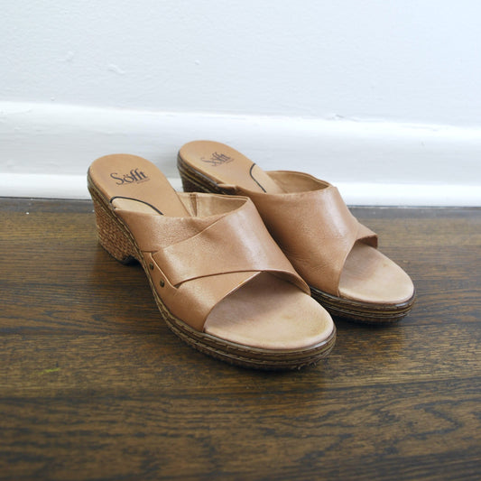Pair of brown sandals on a wooden floor with a white baseboard in the background.