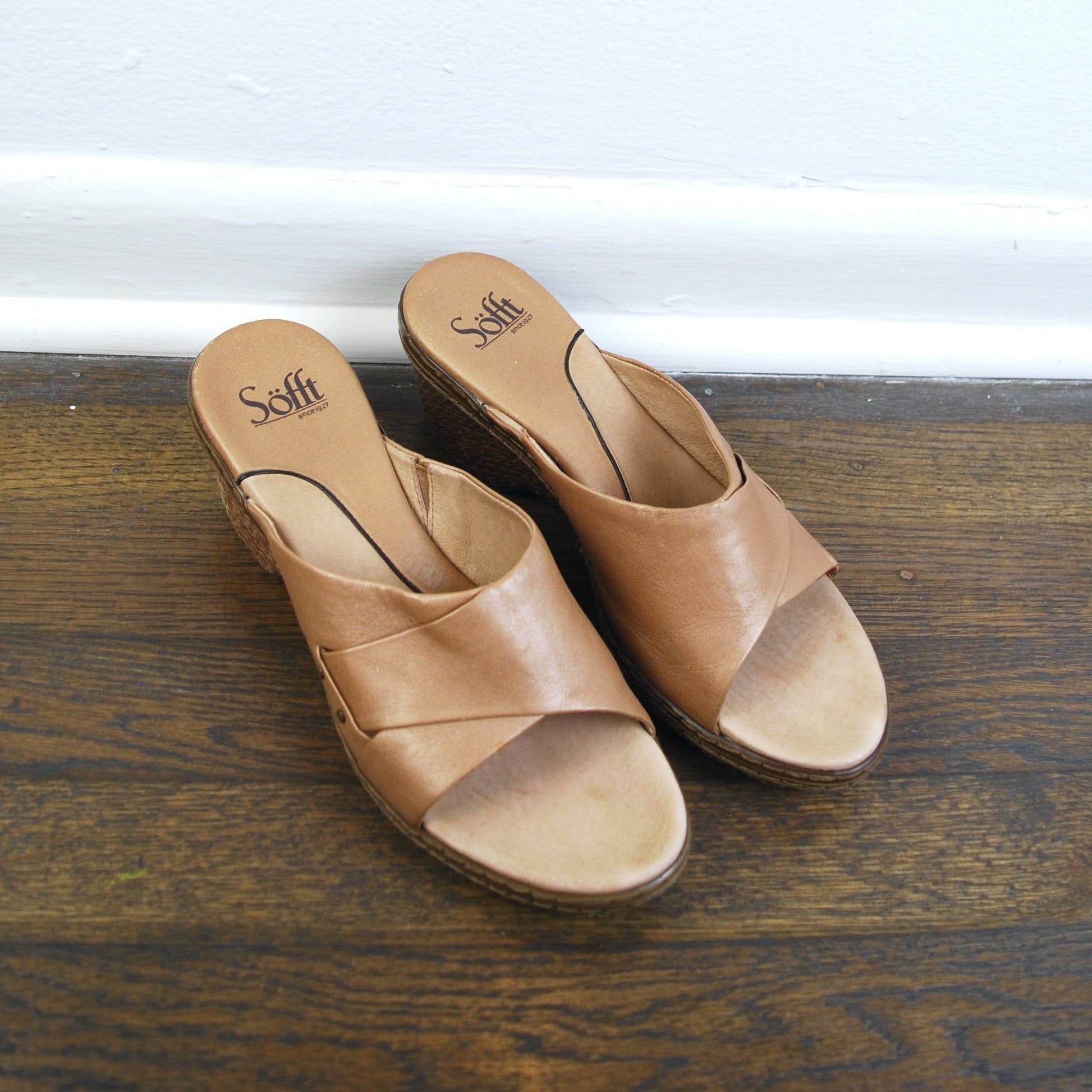Pair of brown sandals on a wooden floor with a white baseboard.