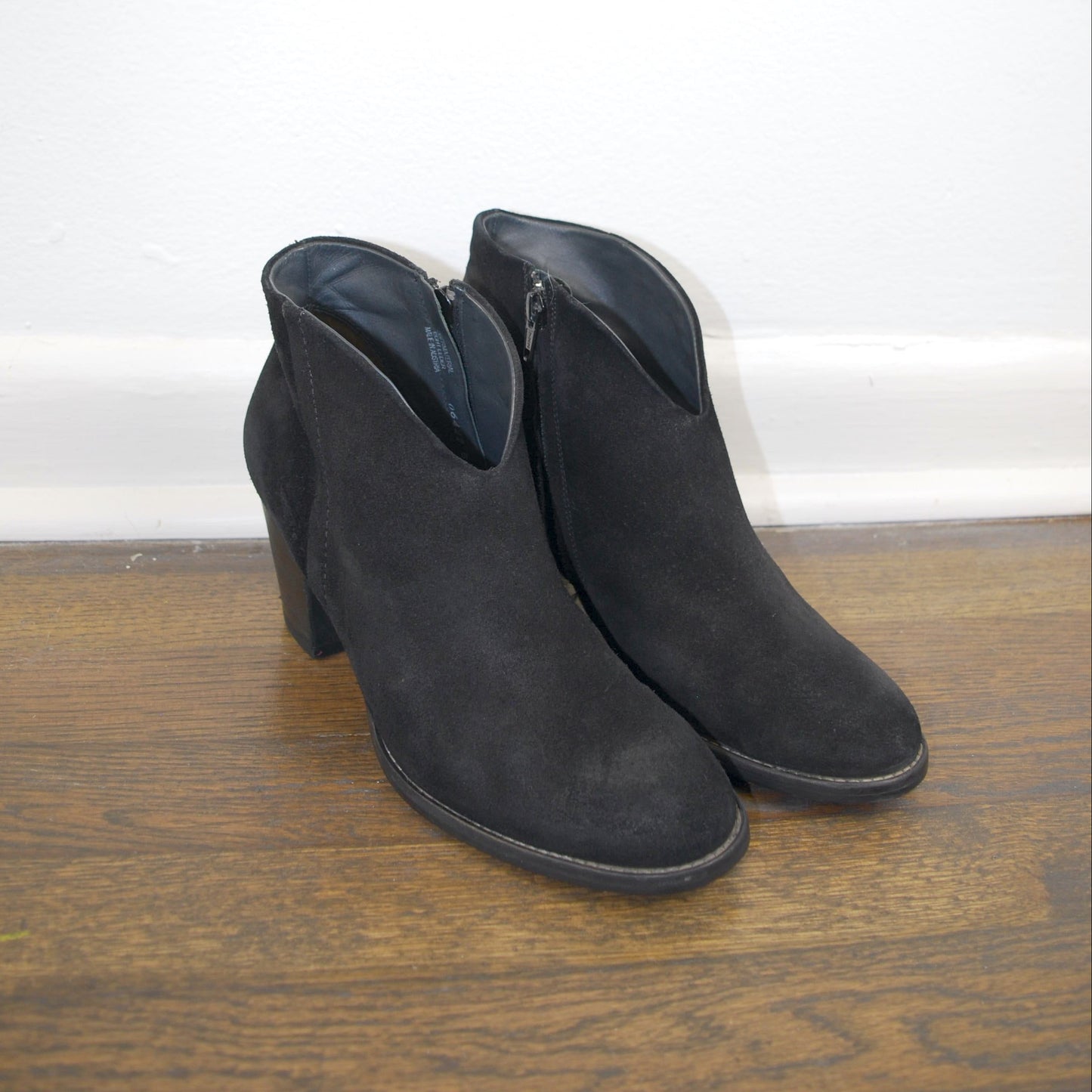 Pair of black suede ankle boots on a wooden floor with a white baseboard.