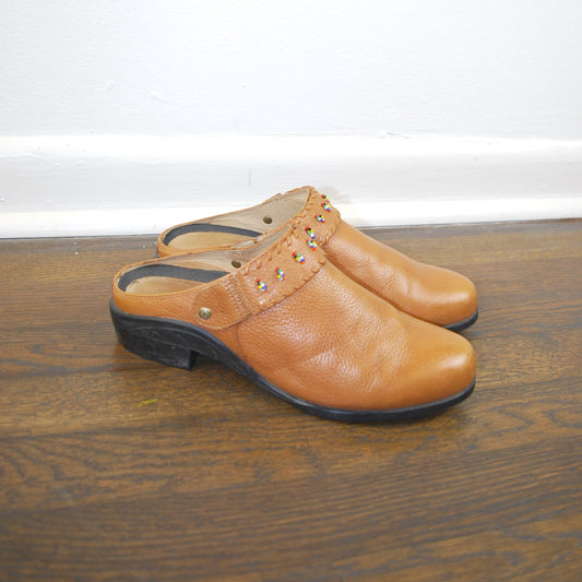 Pair of brown leather clogs on a wooden floor with a white wall background