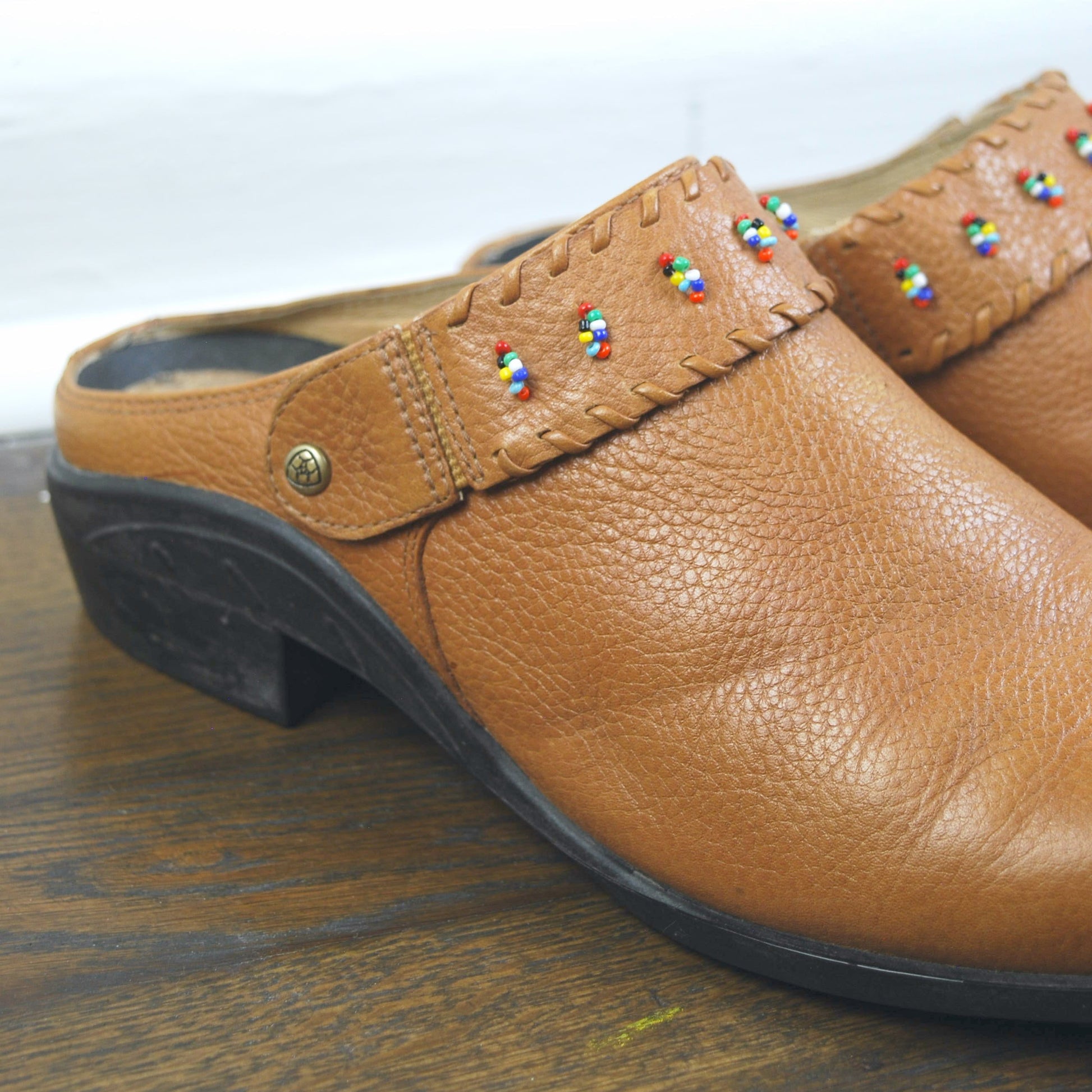 Brown leather clog shoe with colorful embellishments on a wooden surface.