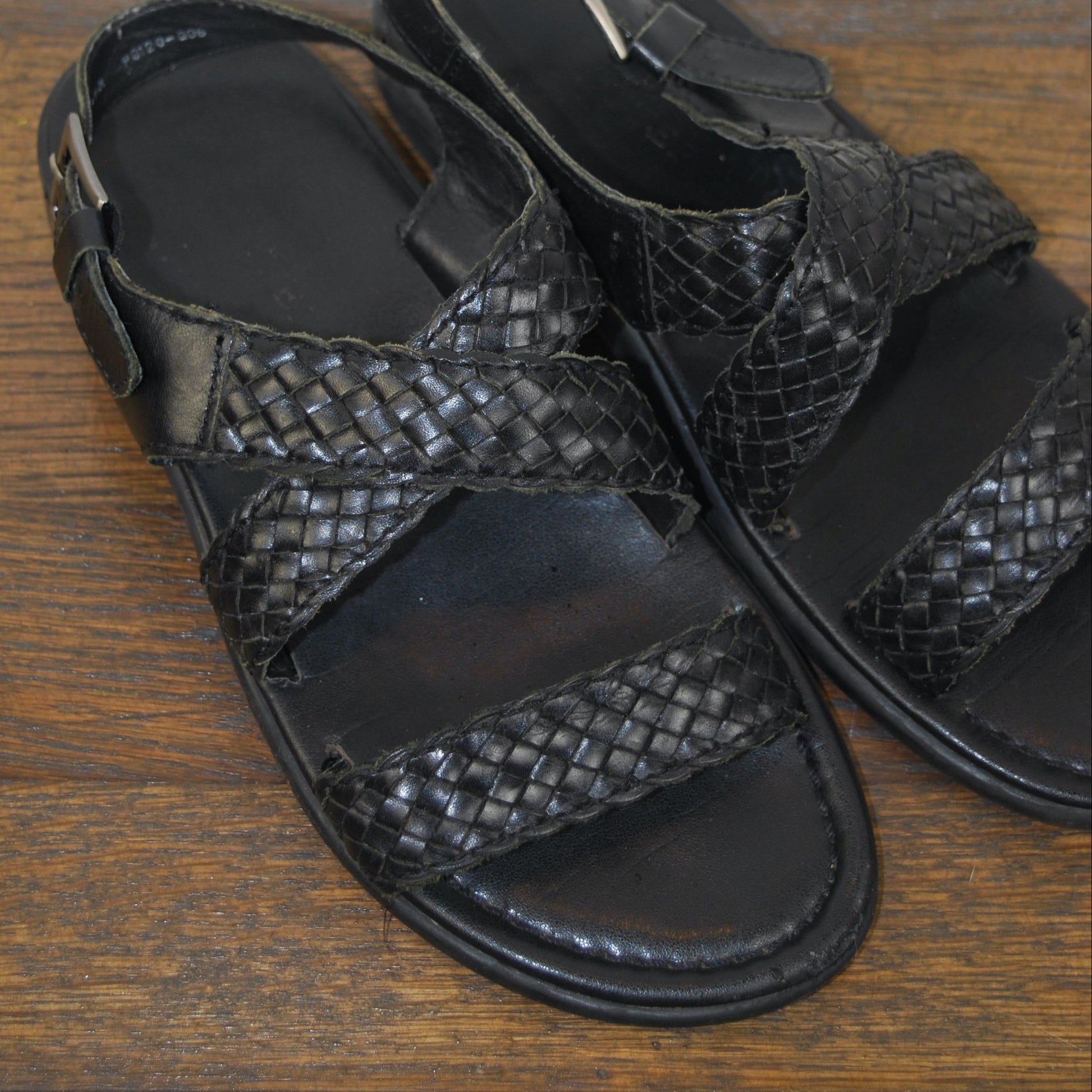 Pair of black sandals on a wooden floor with a white baseboard in the background.